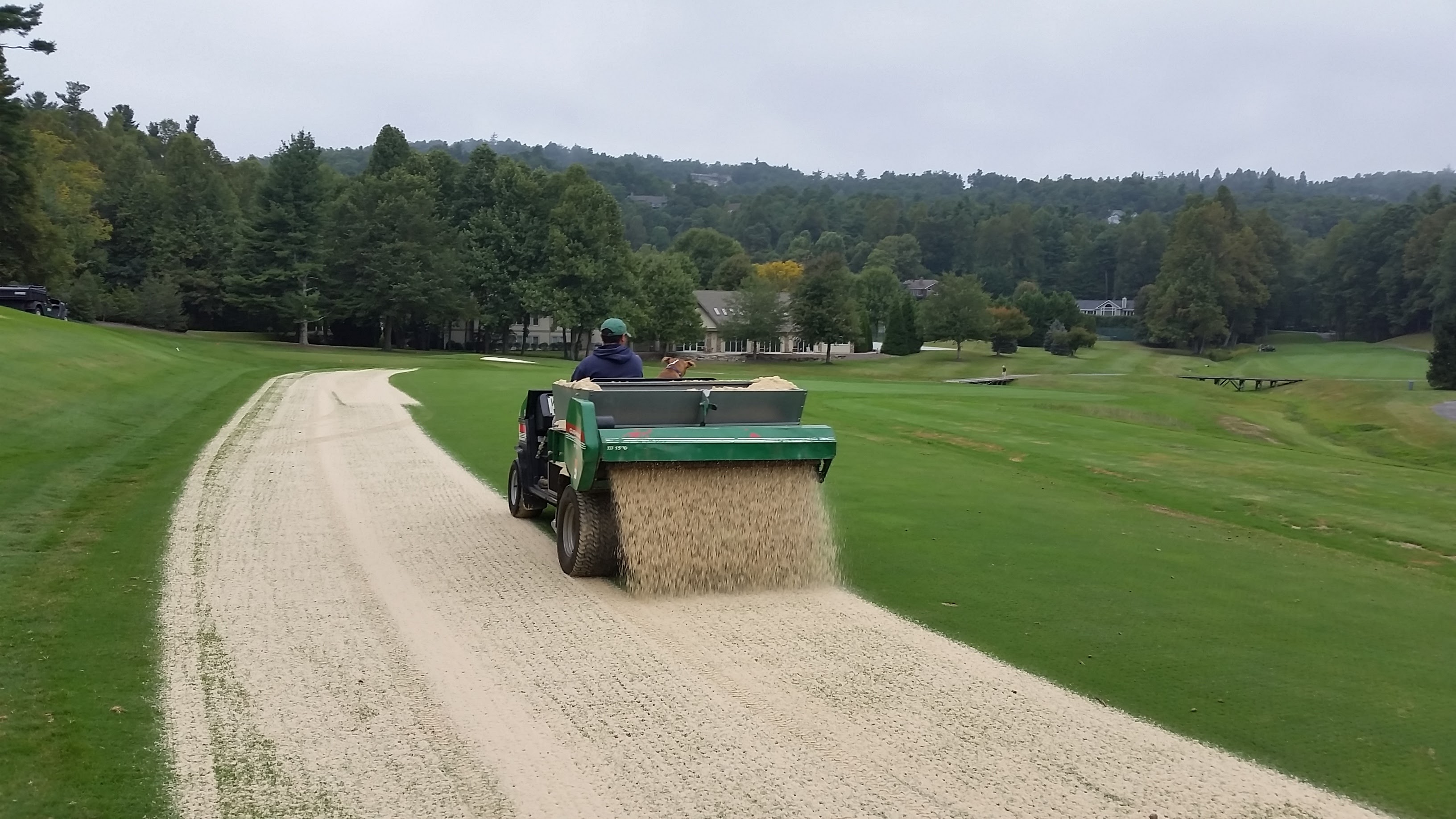 Worker operating a green topdresser spreading sand along a fairway with mountain golf course scenery.