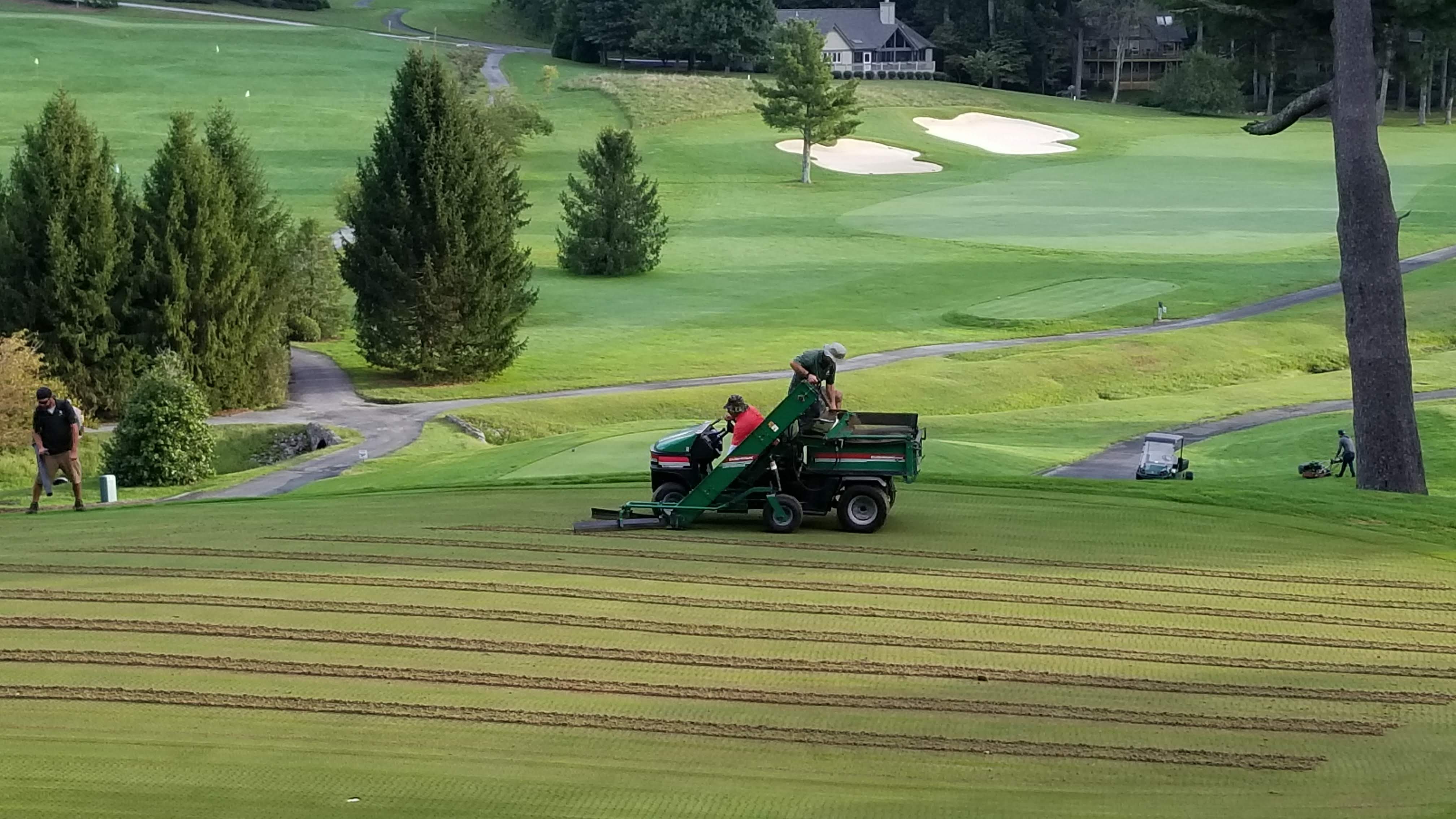 Crew performing greens aerification using a tractor-mounted aerifier with conveyor belt on a sloped green.