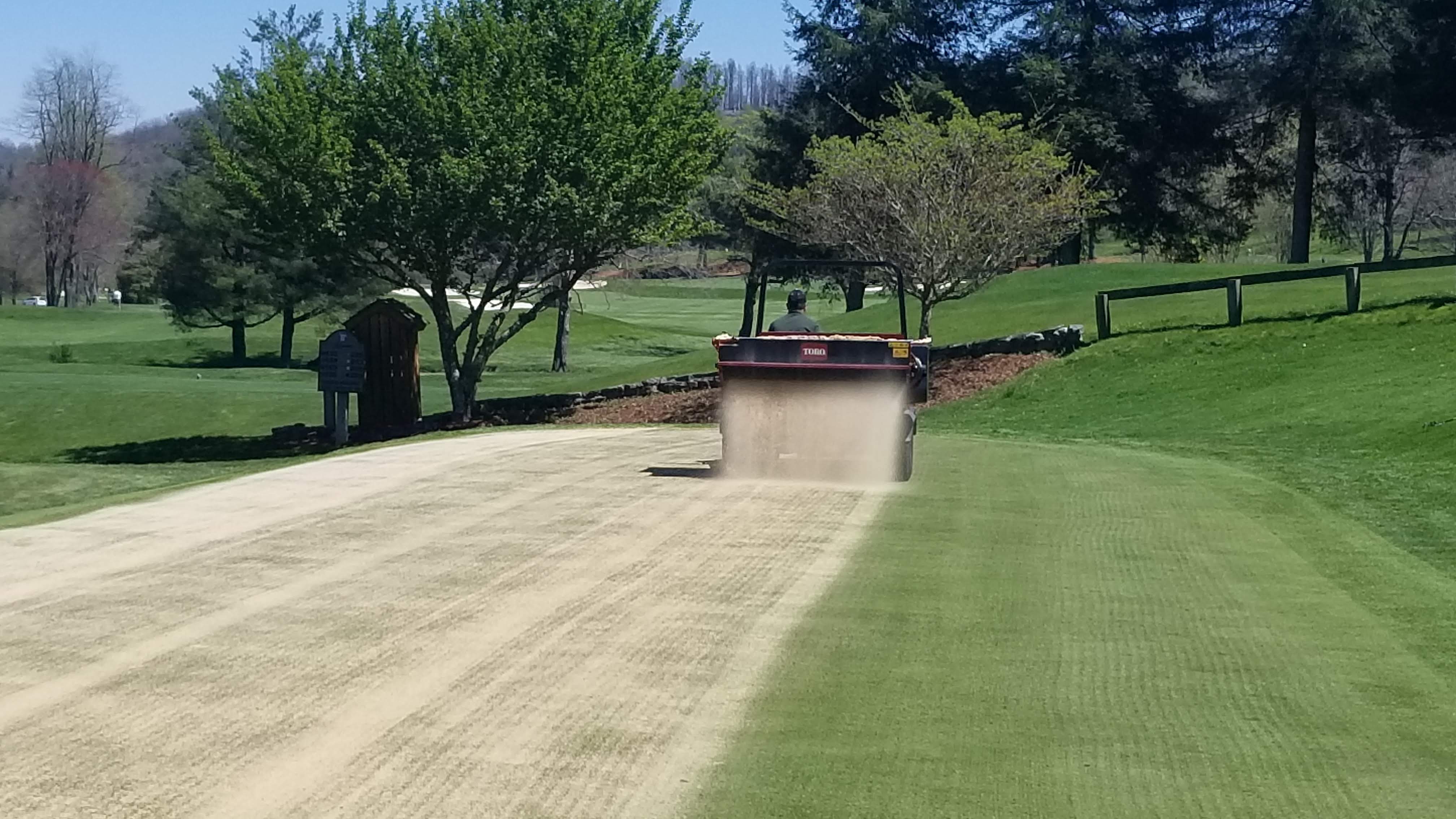 Toro topdresser applying sand to turf with dust rising in early spring conditions.