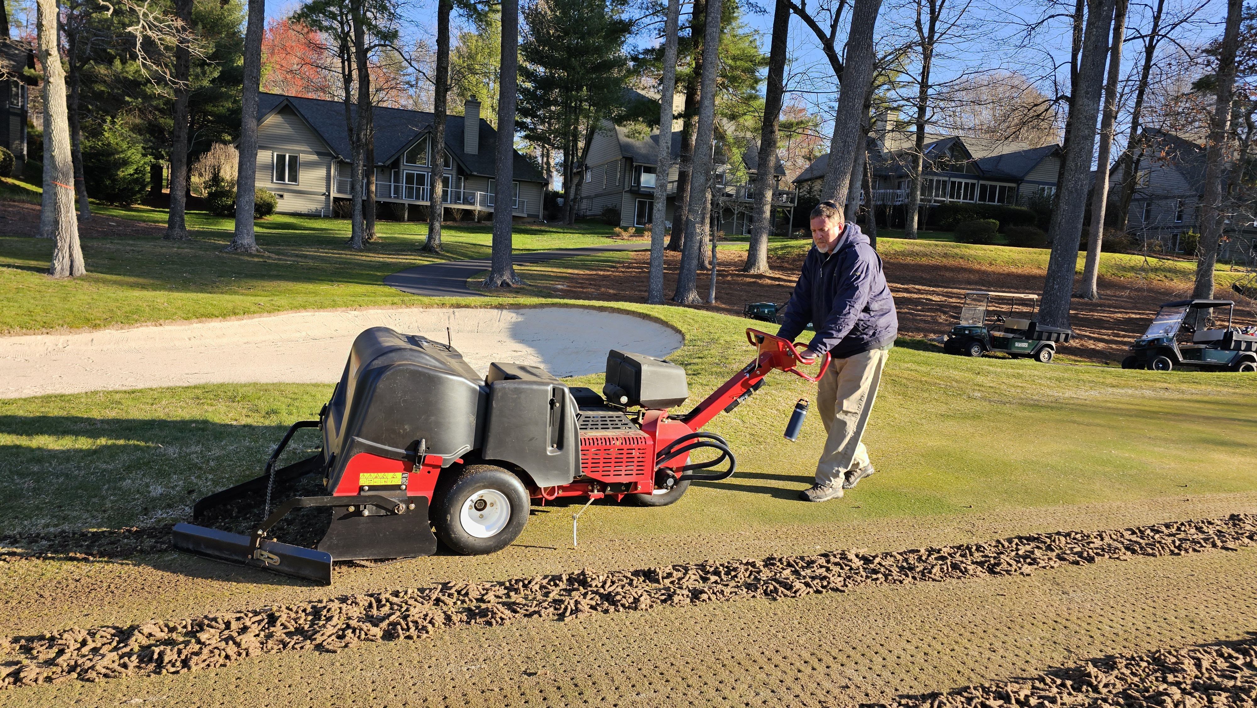 Crew member operating a Toro ProCore aerifier pulling cores from a green while support equipment waits in the background.