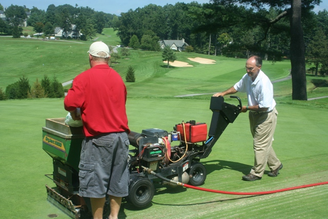 Two turf managers operating a greens aerator with mountainous golf course scenery behind them.