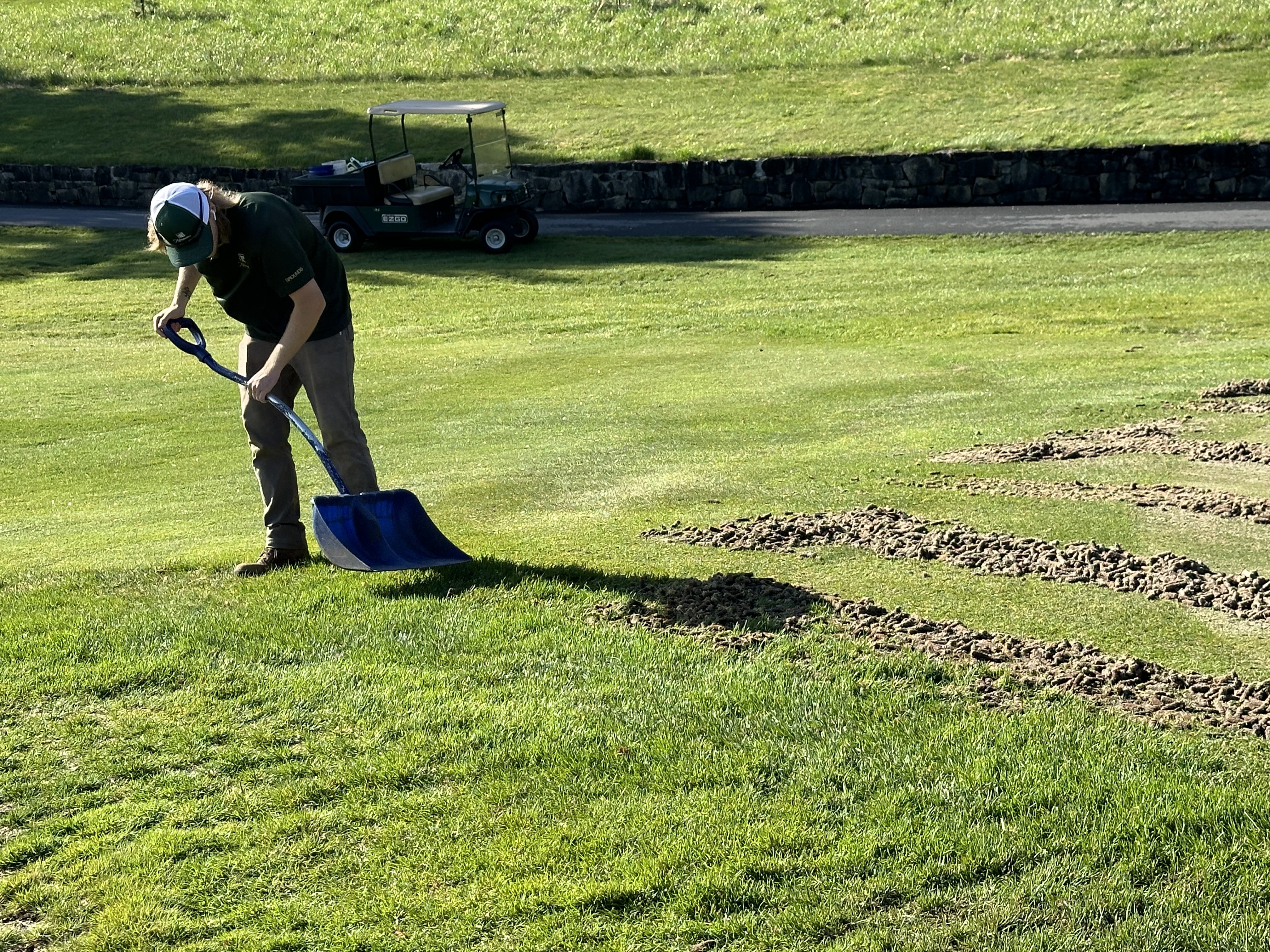 Crew member using a blue scoop shovel to collect soil cores with a utility cart in the background.
