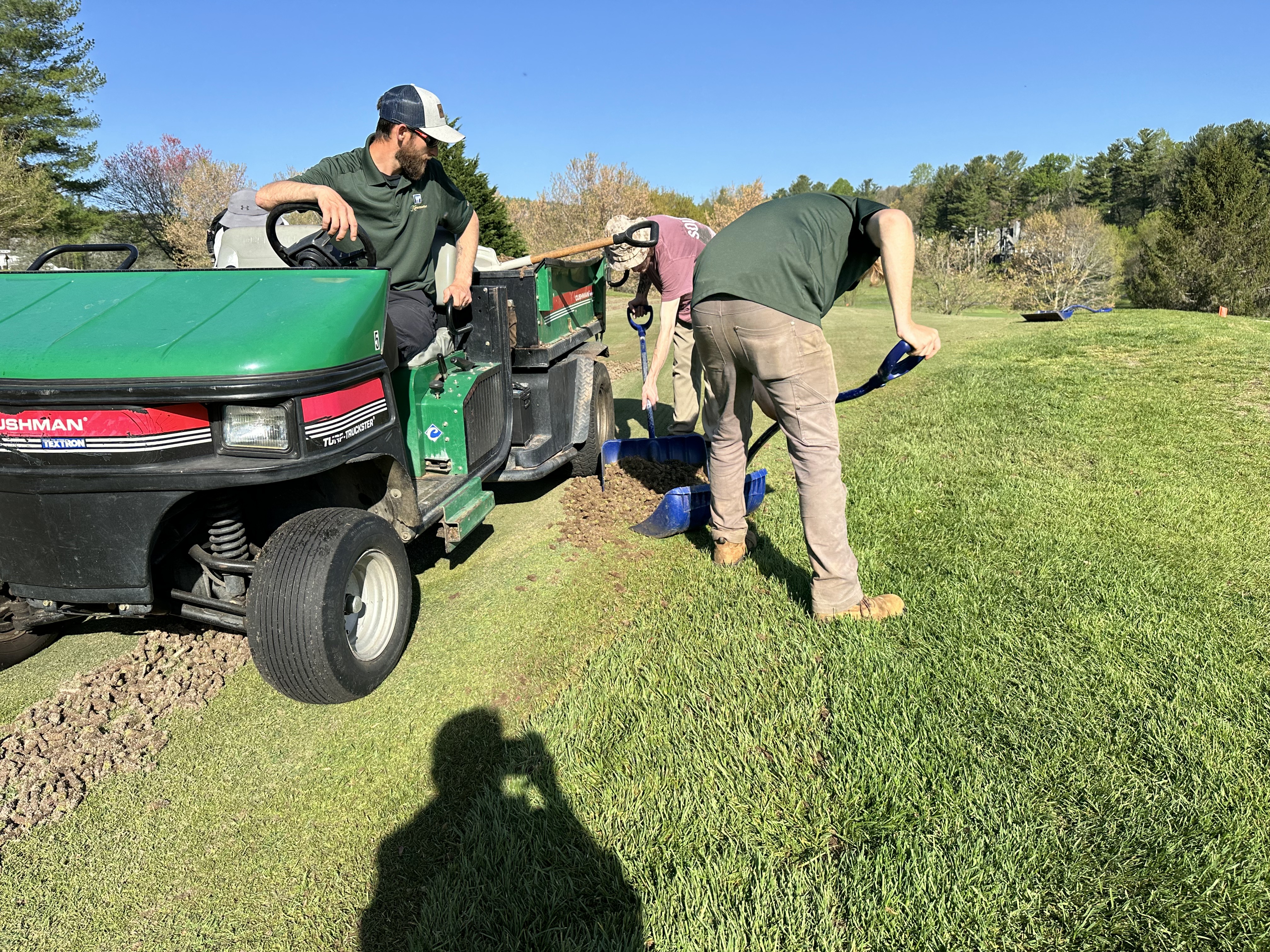 Two workers shoveling aerification cores into a Cushman Turf-Truckster while a third monitors the load.