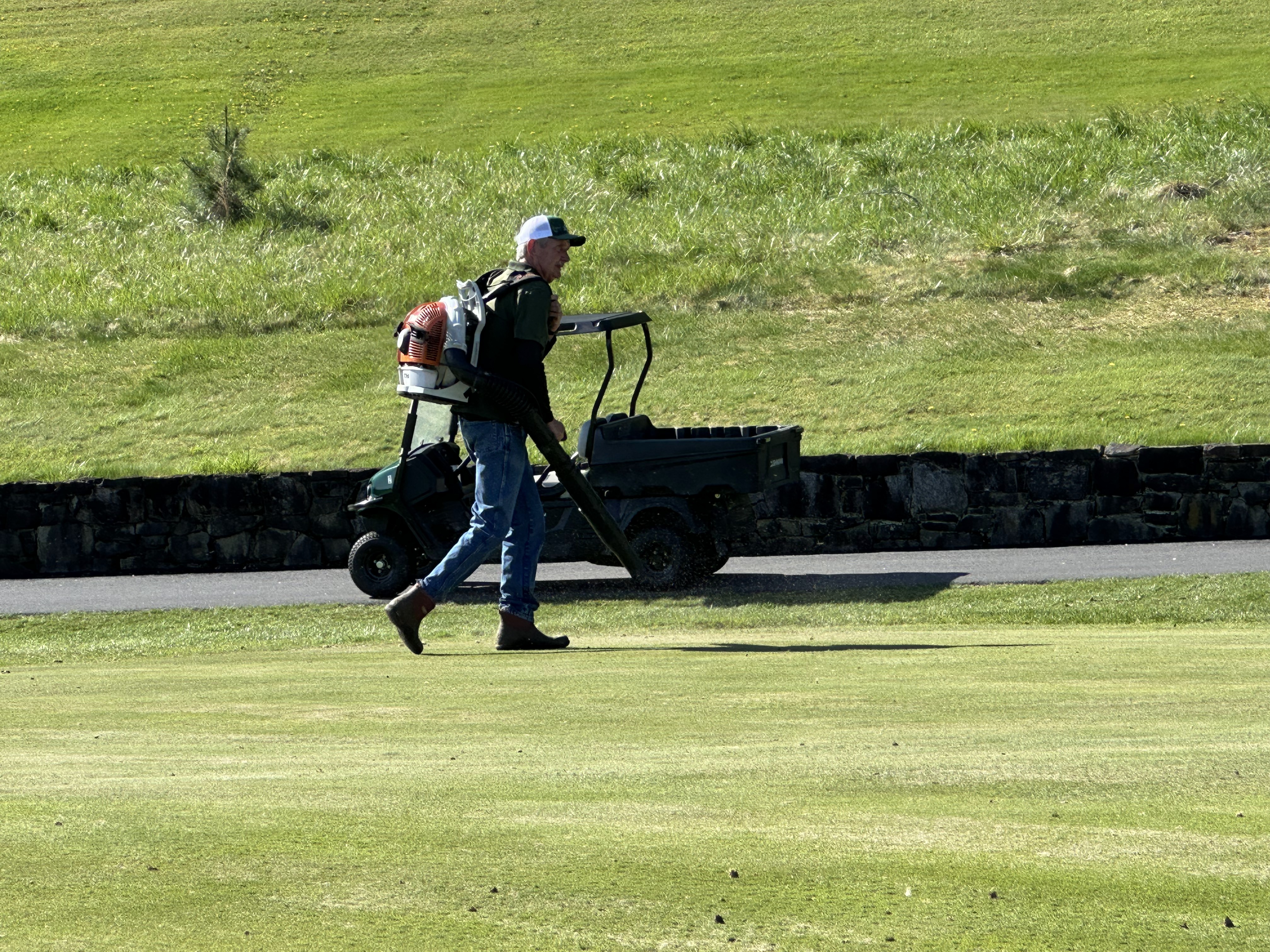 Crew member with a backpack blower clearing debris beside a John Deere utility cart.