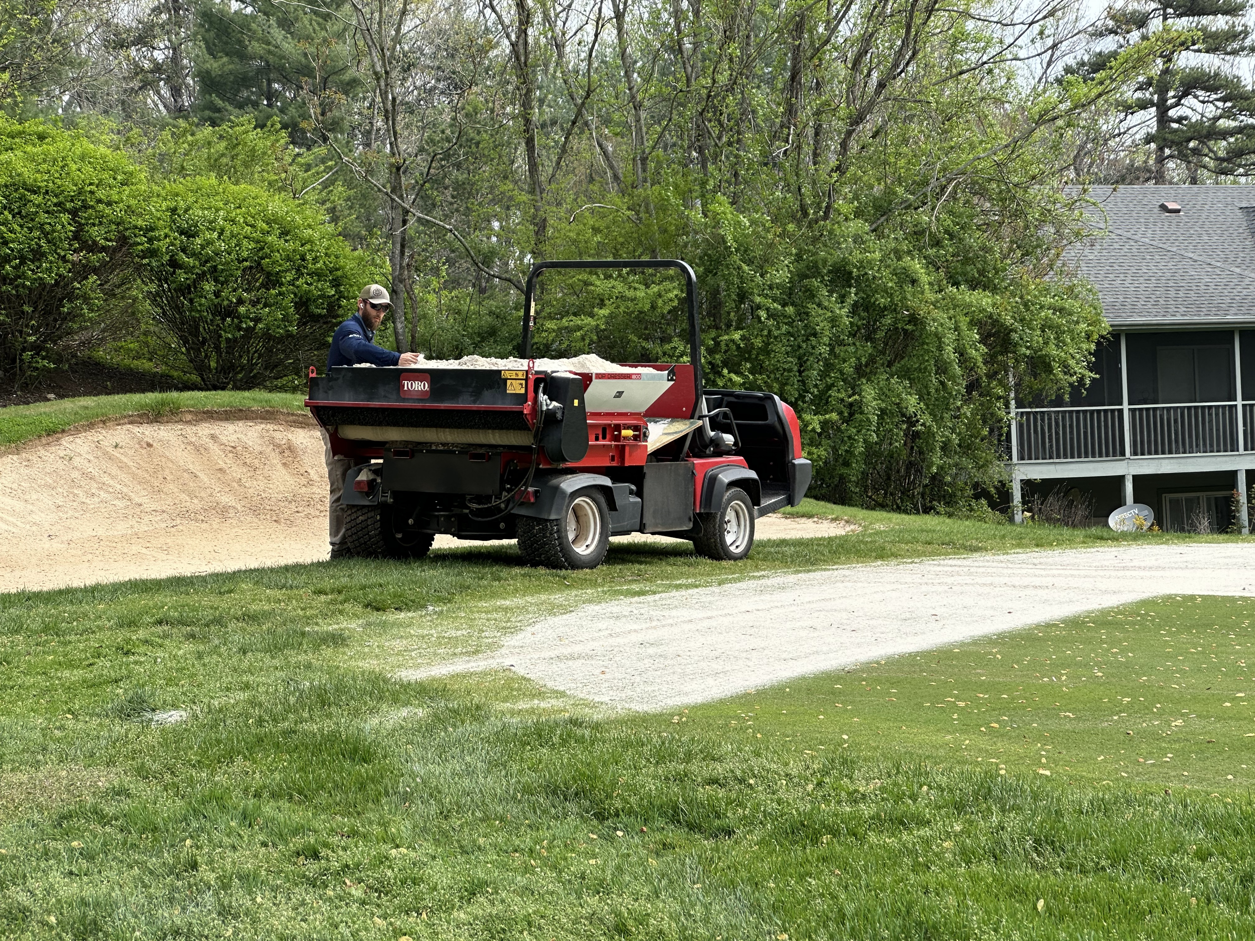 Worker operating a Toro topdresser to apply sand to a bunker near lush shrubs and a clubhouse.